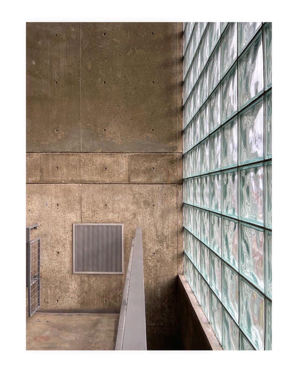 A photograph of a modern architectural interior featuring a wall of textured, exposed concrete panels on the left and a tall, multi-story wall of glass blocks on the right. A metal handrail runs along a concrete ledge in the foreground, with a square ventilation grate set into the concrete wall.