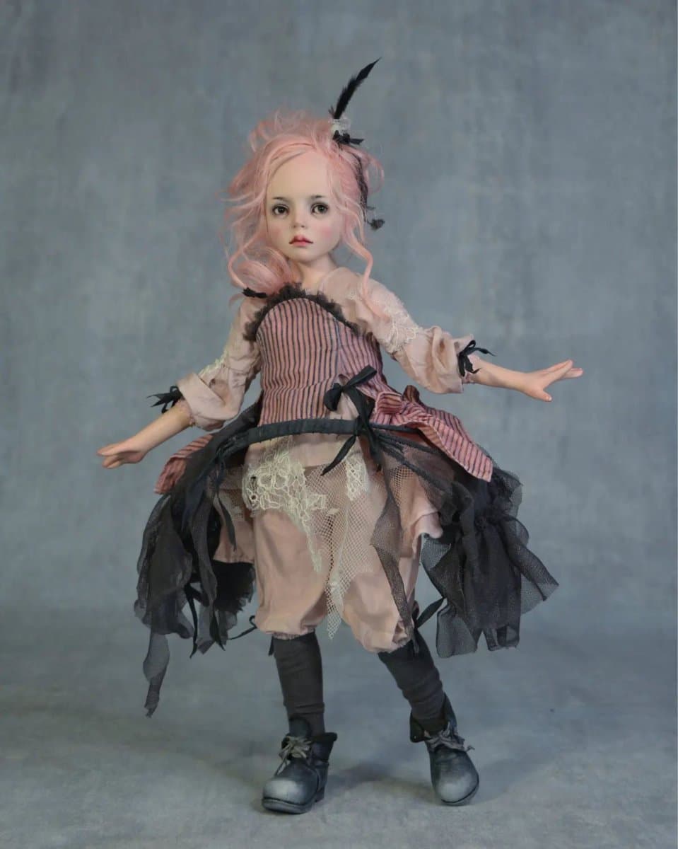 A full-body studio photograph of a handmade doll with pink hair, dressed in a layered pink and black outfit with lace and tulle details, standing against a textured grey background.