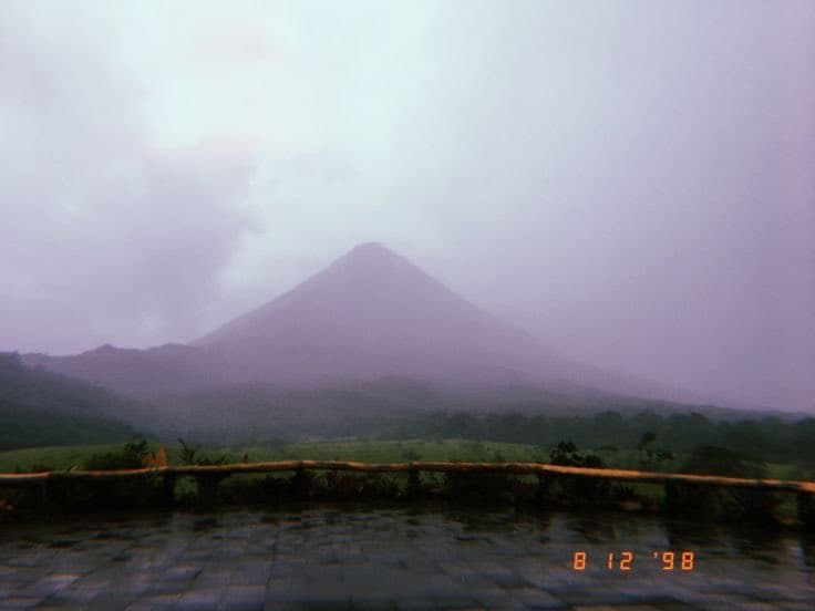 A landscape photograph showing a conical mountain shrouded in mist and clouds, viewed from a wet, tiled terrace with a wooden railing in the foreground.