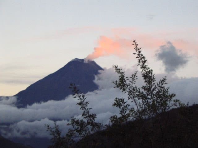 A landscape photograph of a mountain peak emitting a plume of smoke or steam into a pink and blue sky, with a layer of clouds at its base and silhouetted foliage in the foreground.
