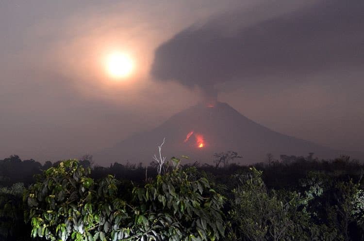 A nighttime photograph of a volcano erupting, with a large plume of dark smoke rising from its peak and glowing lava visible on its slopes. The bright moon shines in the hazy sky above the volcano, which is framed by dark foliage in the foreground.