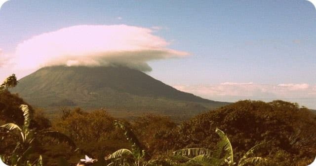 A landscape photograph showing a large, conical volcano with a flat top, partially obscured by a large, flat-bottomed cloud formation hovering directly above its peak. The foreground features lush, green tropical vegetation, including banana leaves, under a bright blue sky.