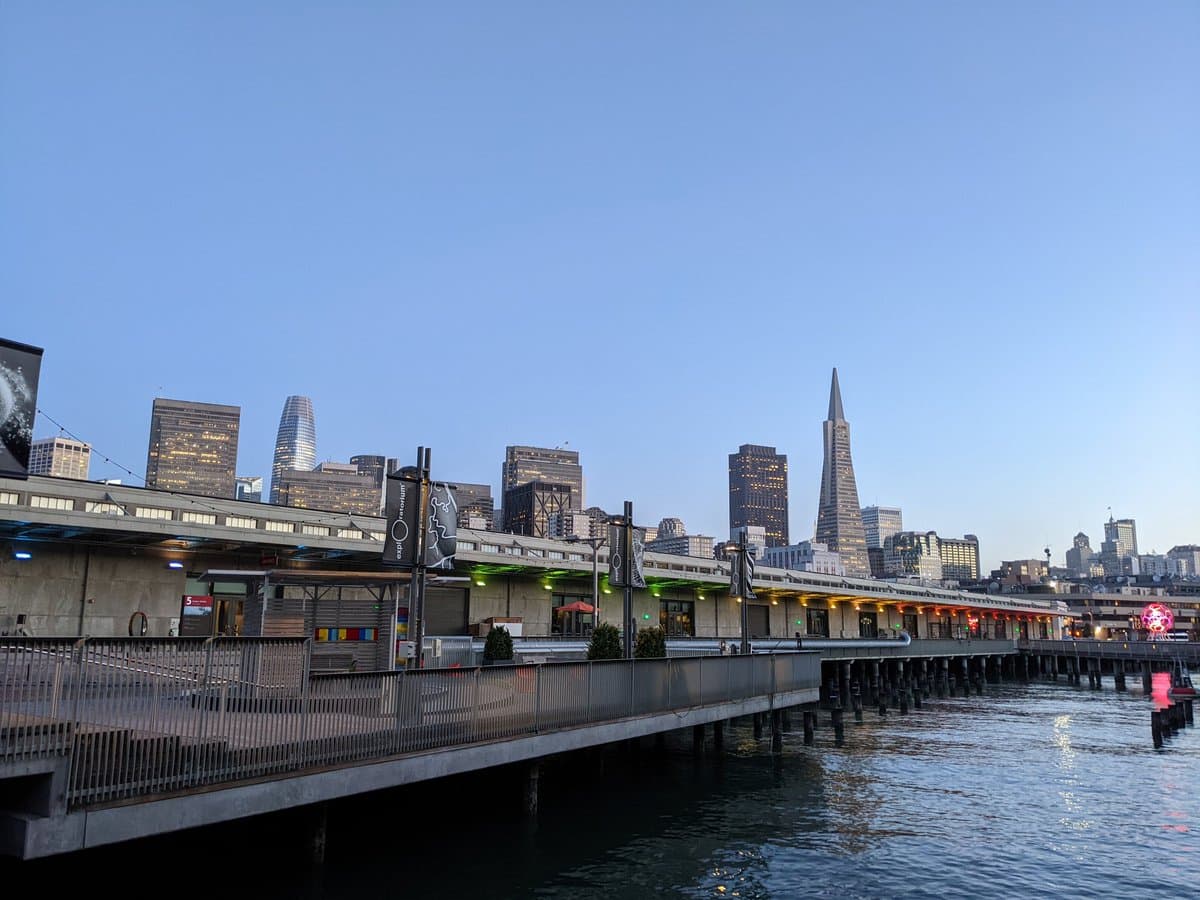 A wide-angle photograph of the San Francisco skyline at dusk, featuring the Transamerica Pyramid and other skyscrapers rising behind the Ferry Building and a waterfront pier.