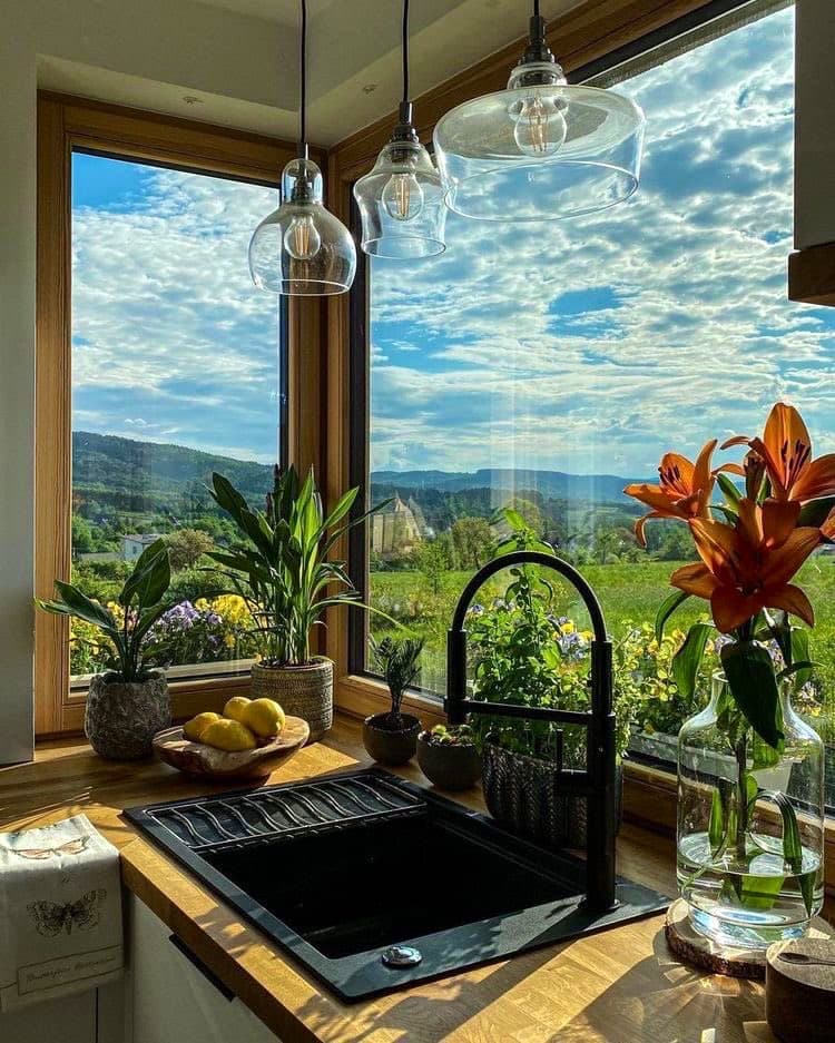 A bright kitchen corner featuring a black farmhouse sink and wooden countertop, set against large windows that reveal a scenic view of green hills and a blue, partly cloudy sky. The space is decorated with hanging glass pendant lights, potted plants, a bowl of lemons, and a vase of orange lilies.