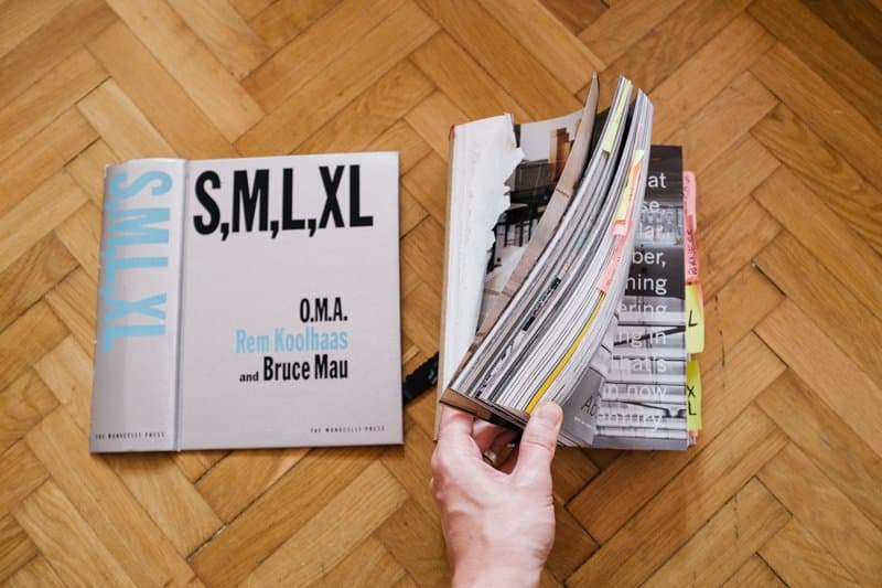 A photograph of a person holding a copy of the book "S,M,L,XL" by Rem Koolhaas and Bruce Mau, with the cover visible on the left and the pages being fanned out on the right, all set on a wooden parquet floor.