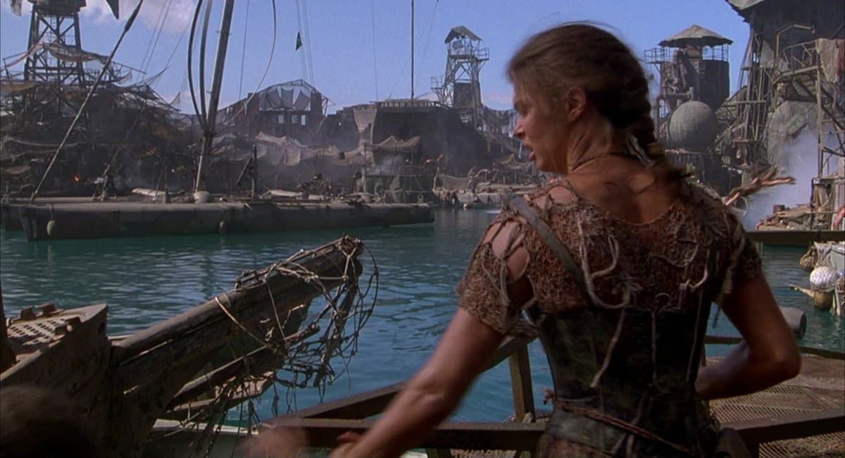 A still from the 1995 film 'Waterworld' showing a woman in a tattered, rope-adorned outfit standing on a wooden boat, looking out over a large, makeshift floating city built from scrap metal and debris on the water.
