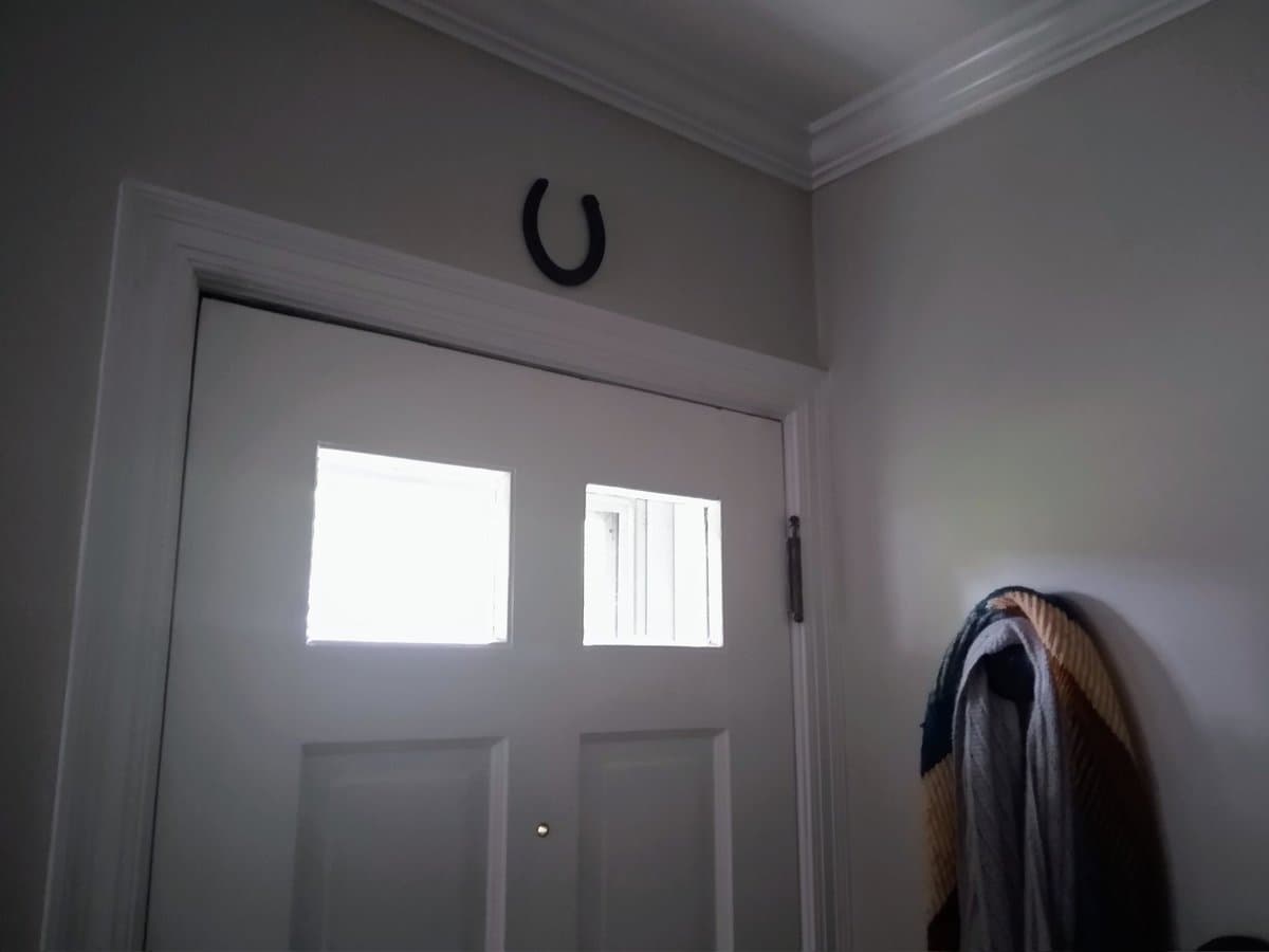 A photo of a white interior door with two windows, featuring a black horseshoe mounted on the wall above the door frame. A coat hangs on a hook to the right of the door.