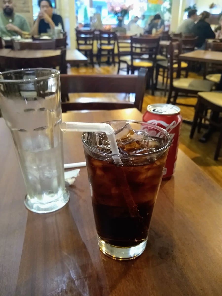 A photograph taken in a restaurant showing a glass of iced coffee with a plastic straw, a glass of water, and a can of Coca-Cola on a wooden table.