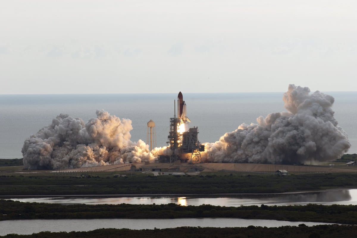 A wide-angle photograph of a space shuttle launching from a coastal launchpad, with massive plumes of white smoke billowing from the base of the vehicle. The launch site is situated on a grassy peninsula surrounded by water, with the ocean visible in the background under a clear sky.