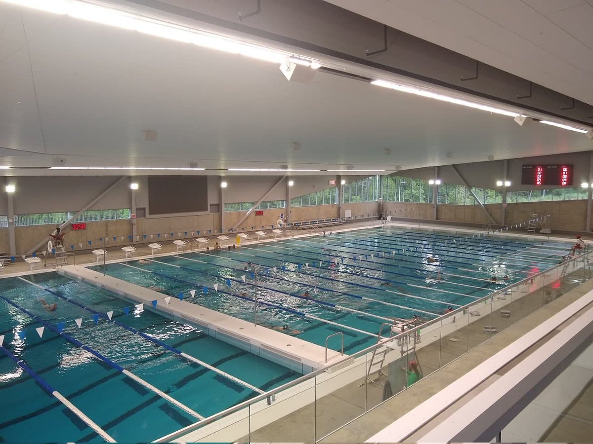 A wide-angle photograph of a large indoor swimming facility featuring two pools with multiple lanes and swimmers. The scene is illuminated by bright overhead lights and natural light from a row of windows, with digital scoreboards visible on the far wall.