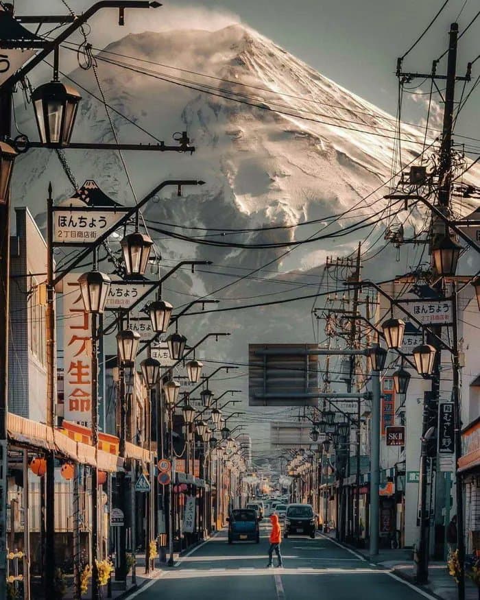 A street-level photograph looking down a narrow Japanese shopping street lined with vintage-style street lamps and storefronts, with the snow-capped peak of Mount Fuji towering in the background. A person in an orange jacket walks in the middle of the road, and a blue car is parked nearby. The scene is framed by overhead power lines and Japanese signage on the buildings.