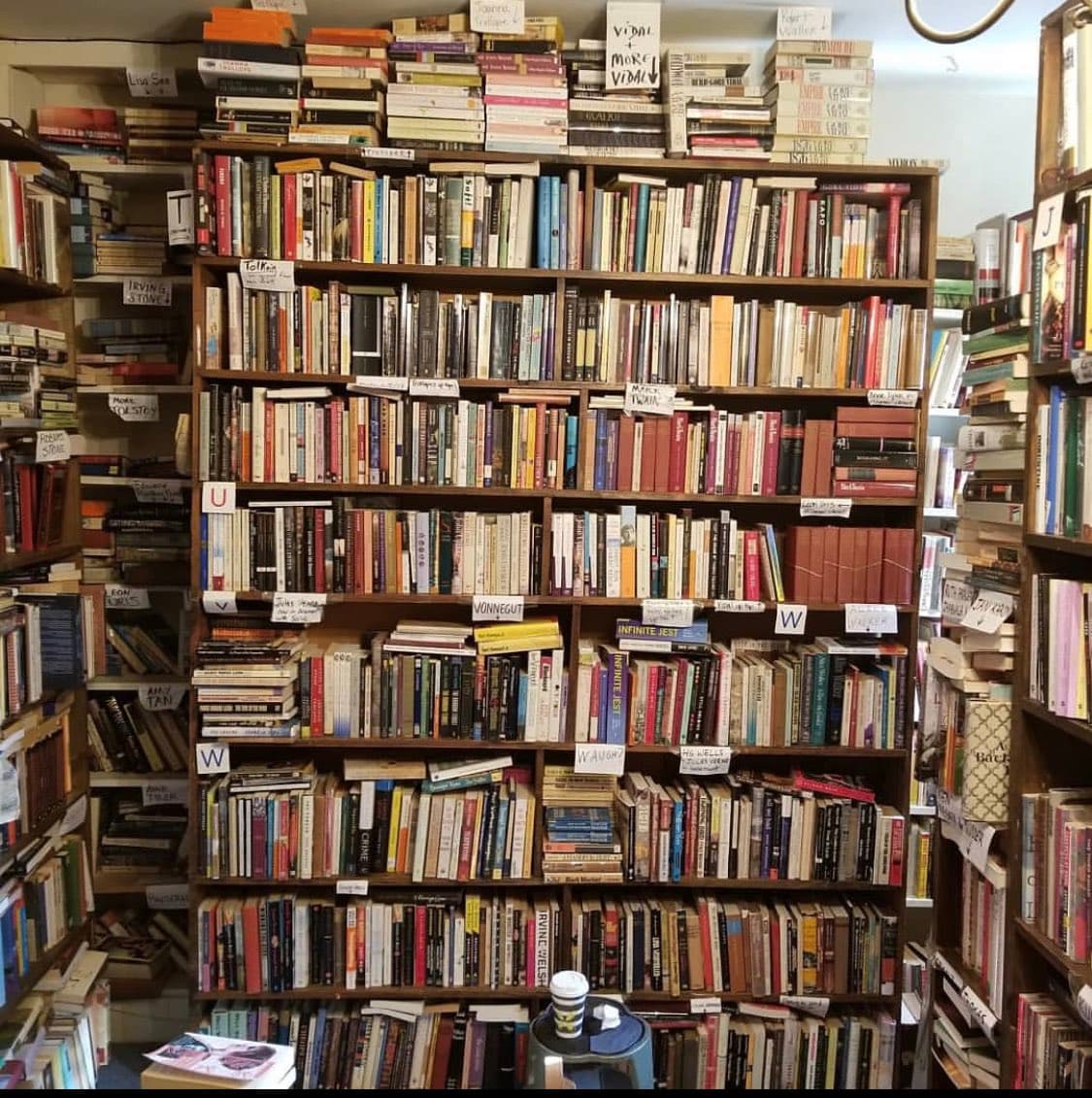 A photograph of a densely packed bookshelf in a bookstore, filled with numerous books and handwritten author name labels. A small table with a coffee cup sits in the foreground.