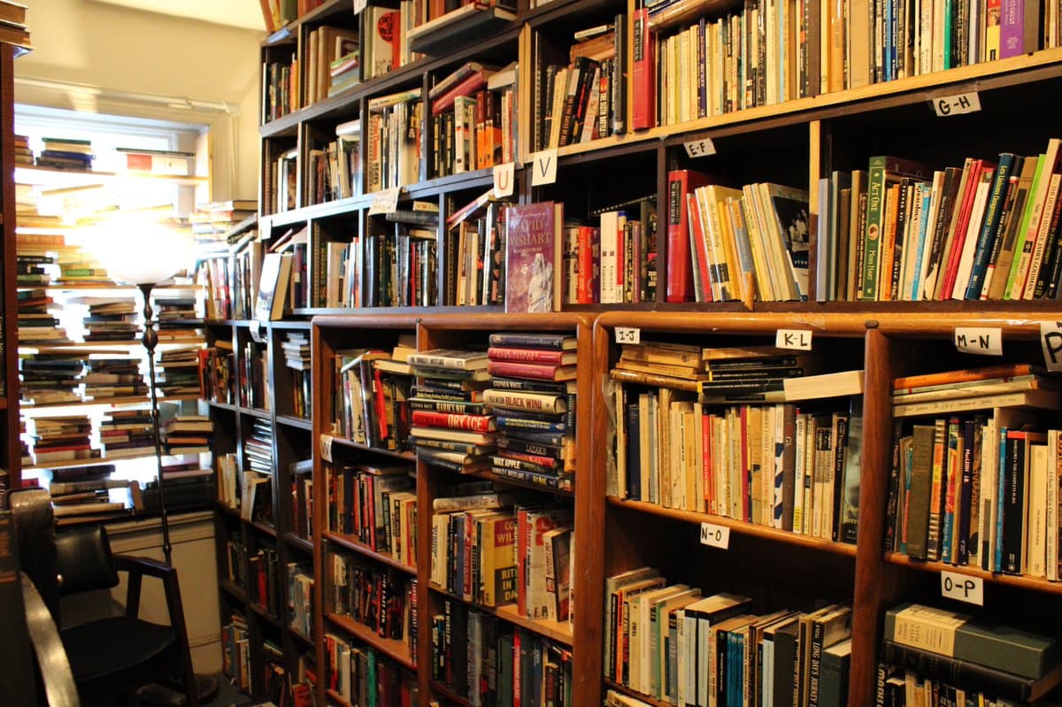 A photograph of a cozy bookstore interior featuring wooden bookshelves packed with books and a floor lamp near a window. The shelves are organized with handwritten paper labels indicating alphabetical sections.