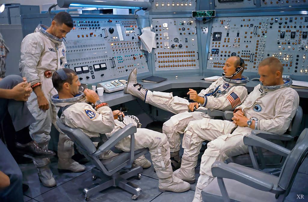 A photograph showing four astronauts in white spacesuits inside a control room with complex instrument panels, where three are seated in chairs and one is standing.