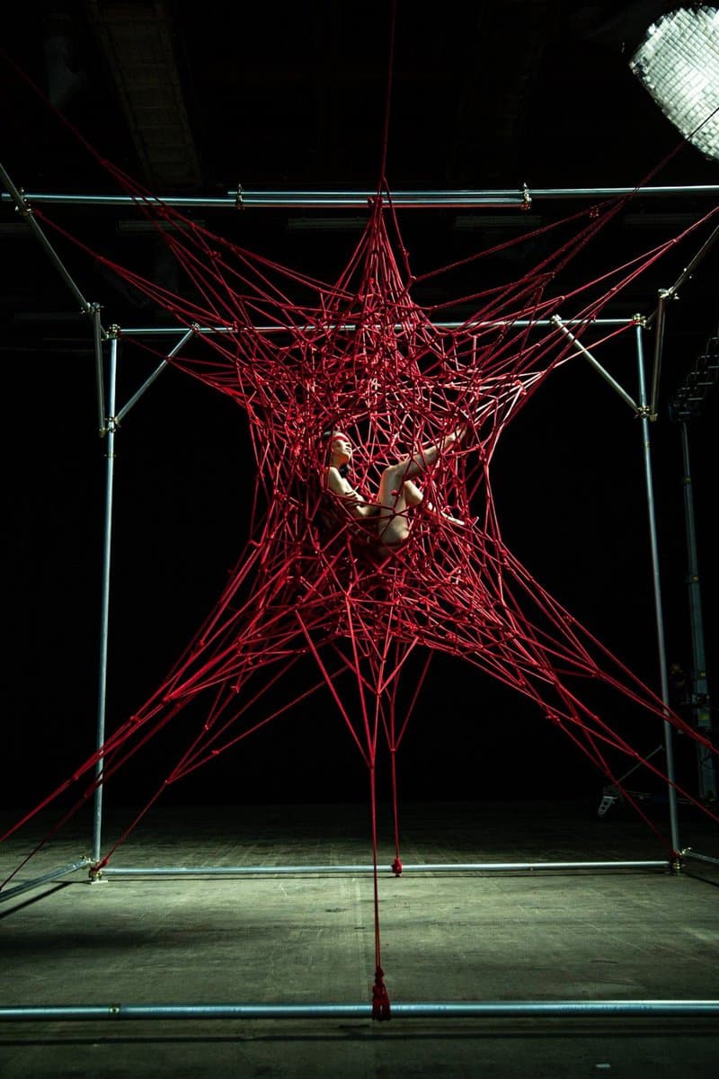 A person is suspended in the center of a large, star-shaped structure made of red rope, which is stretched across a metal scaffolding frame in a dark, industrial space.