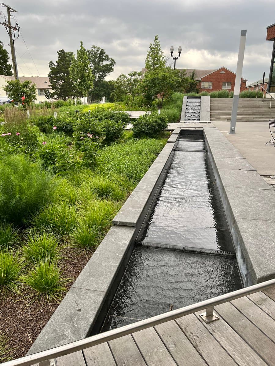 A biophilic water feature with a long, rectangular stone channel filled with flowing water, situated next to a landscaped garden with green plants and flowers. In the background, there is a paved plaza, a small waterfall, and residential buildings under a cloudy sky.