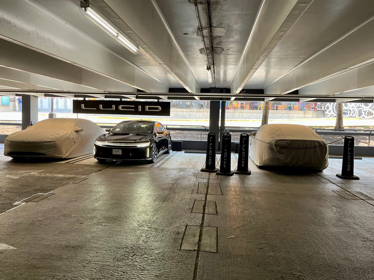 A photo of a Lucid Motors display area in a parking garage, featuring a black electric sedan parked between two cars covered in beige protective sheets. A large black sign with the white "LUCID" logo hangs above the vehicles, and several black charging stations are positioned nearby.