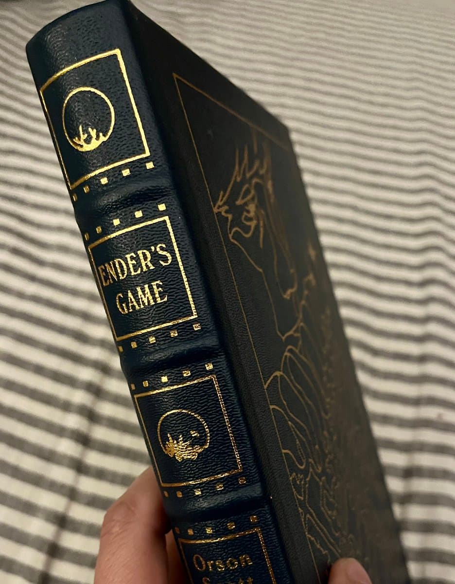 A close-up photograph of a black, leather-bound hardcover book held in a person's hand, showing the spine and part of the front cover. The spine features gold-embossed text and decorative elements, including the title and author's name.