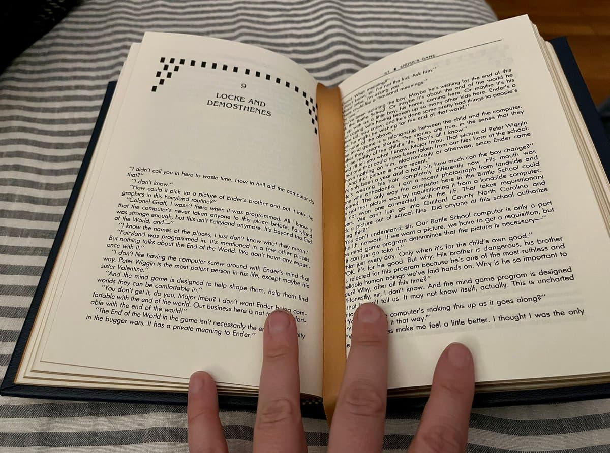 A photograph of an open hardcover copy of the novel 'Ender's Game' resting on a striped fabric surface, with a person's hands holding the pages open. The left page displays the chapter title '9 LOCKE AND DEMOSTHENES' and the beginning of the chapter's text.