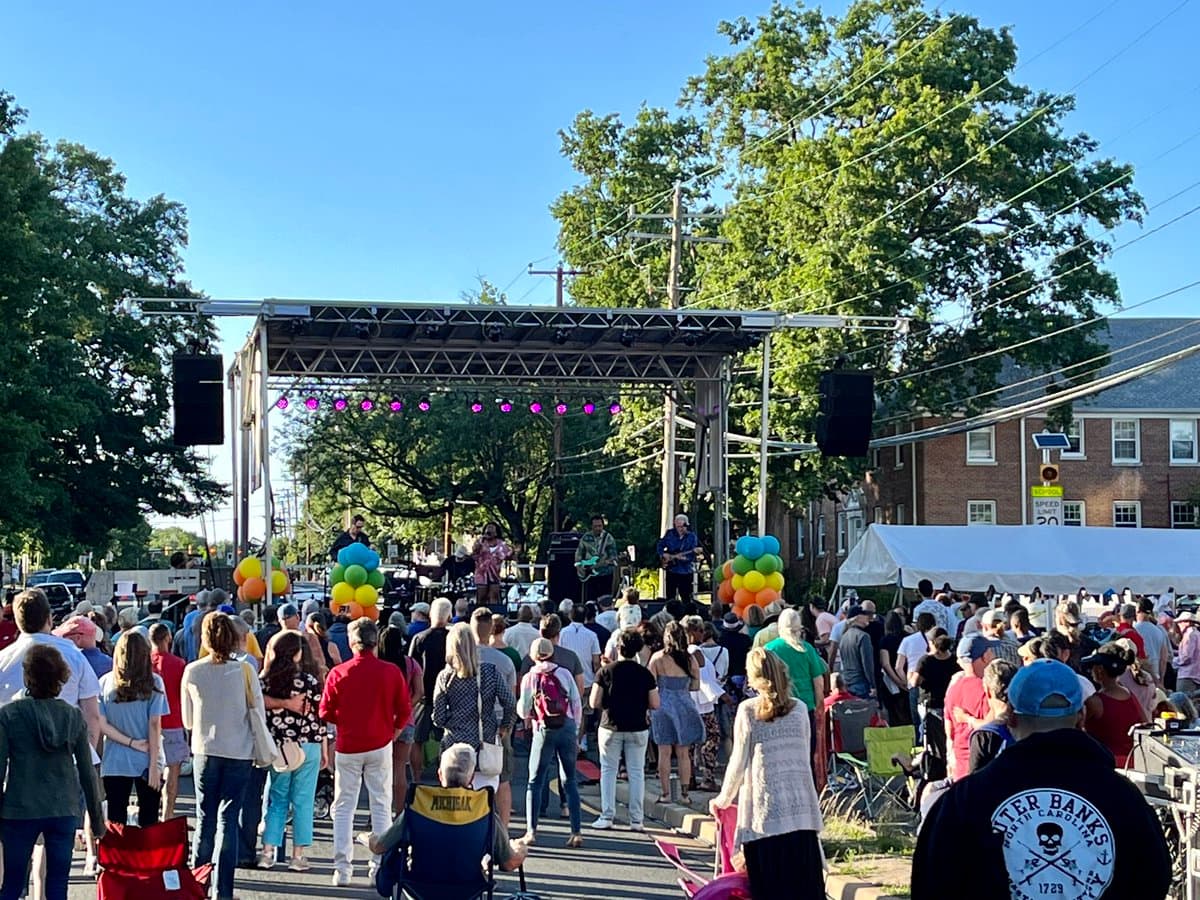 An outdoor photograph showing a large crowd of people gathered on a street to watch a band perform on a stage under a clear blue sky. The scene includes a white tent, trees, and a brick building in the background, with colorful balloons decorating the stage area.
