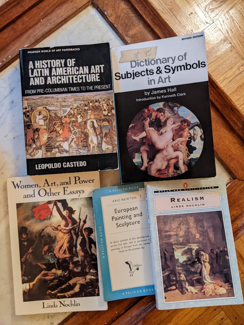 A photograph of five art history books arranged on a wooden surface. The books include titles on Latin American art, art symbols, women in art, European painting, and realism.