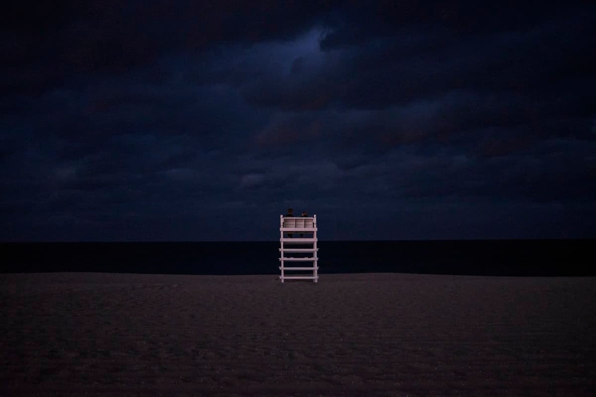 A dark, atmospheric photograph of a beach at night featuring a lone white lifeguard chair in the center against a backdrop of a dark ocean and cloudy sky.