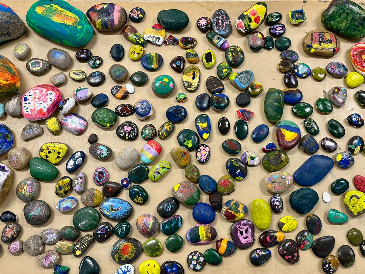A collection of small, smooth river rocks painted with various colorful designs, symbols, and words, arranged on a flat surface.