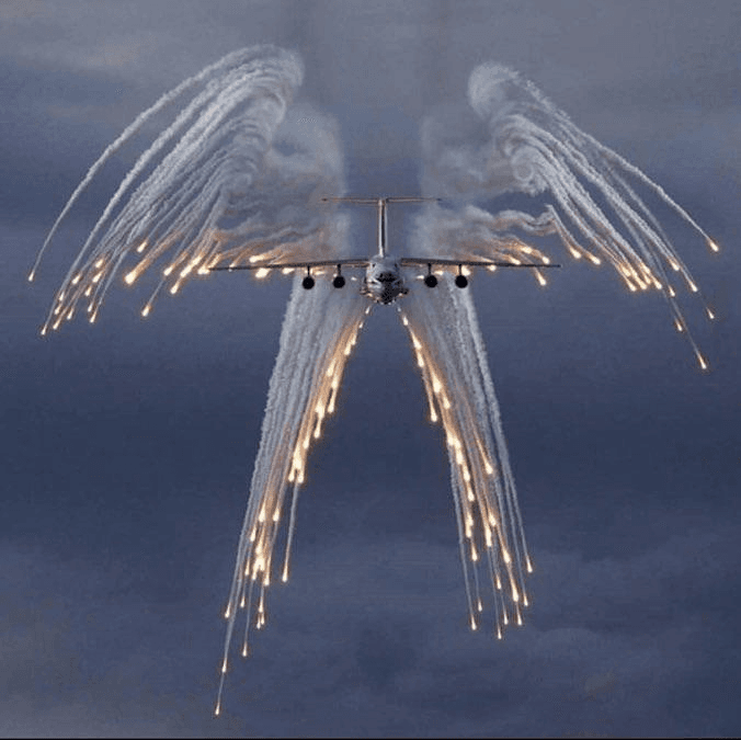 A military aircraft flies against a cloudy sky, trailing smoke and flares from its wings and fuselage that form the shape of angel wings. The image is a photograph used as a visual pun for a 'biblically accurate angel.'