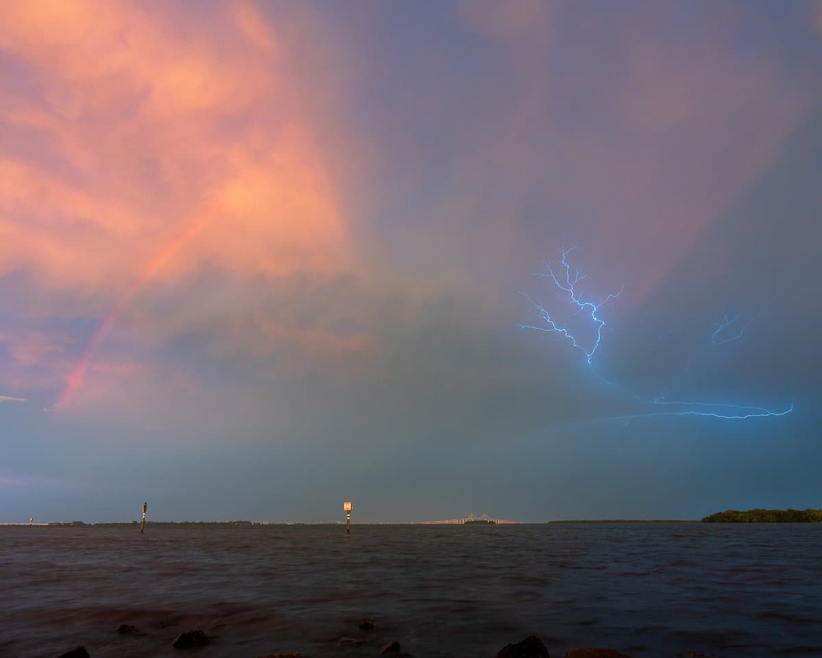 A landscape photograph capturing a dramatic sky over a body of water, featuring a bright rainbow on the left and a branching lightning bolt on the right against a backdrop of pink and purple clouds.