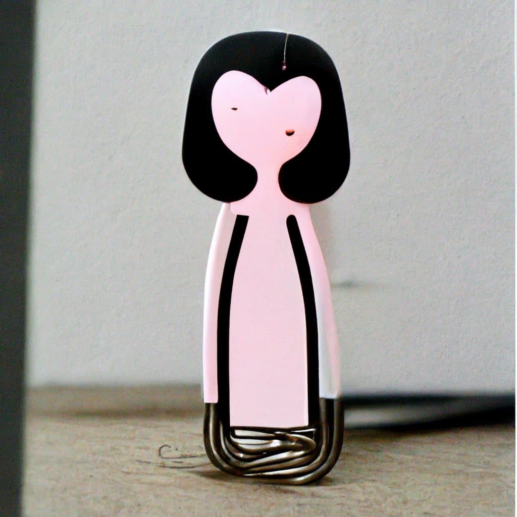 A small, stylized figurine of a person with black hair and a pink body, standing on a metal spring base. The figure has a simple, cartoon-like face with closed eyes and a small mouth.