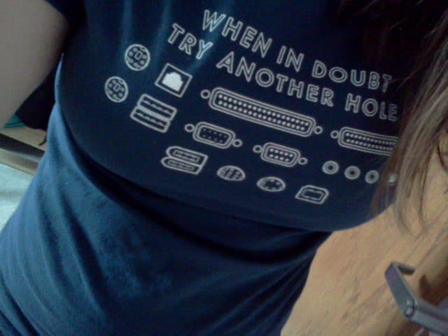 A close-up photograph of a person wearing a dark blue t-shirt featuring white line-art illustrations of various computer ports and connectors. The shirt includes the text "WHEN IN DOUBT TRY ANOTHER HOLE" printed above the graphics.