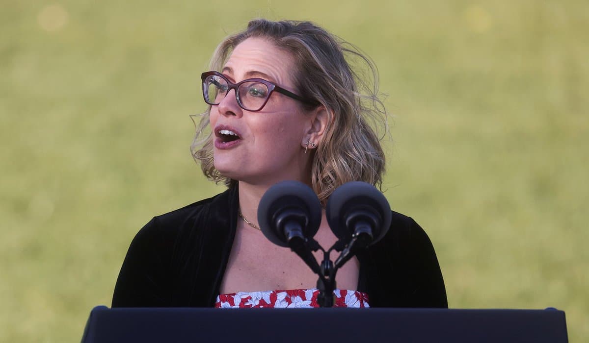 A woman with short blonde hair and glasses stands at a podium with two microphones, speaking outdoors against a blurred green background. She is wearing a black jacket over a red and white floral top.