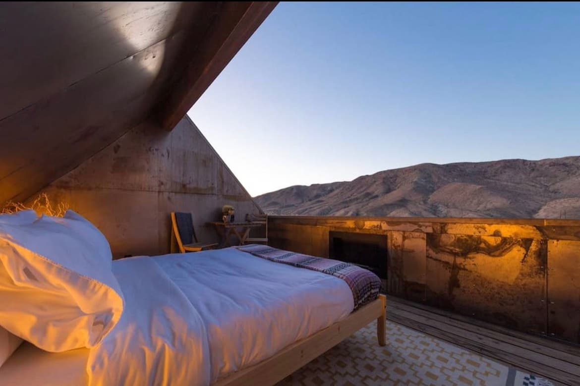 A photograph of an open-air bedroom with a bed, chair, and fireplace set against a backdrop of desert mountains at dusk. The room features concrete walls and a wooden deck floor, illuminated by warm artificial lighting.