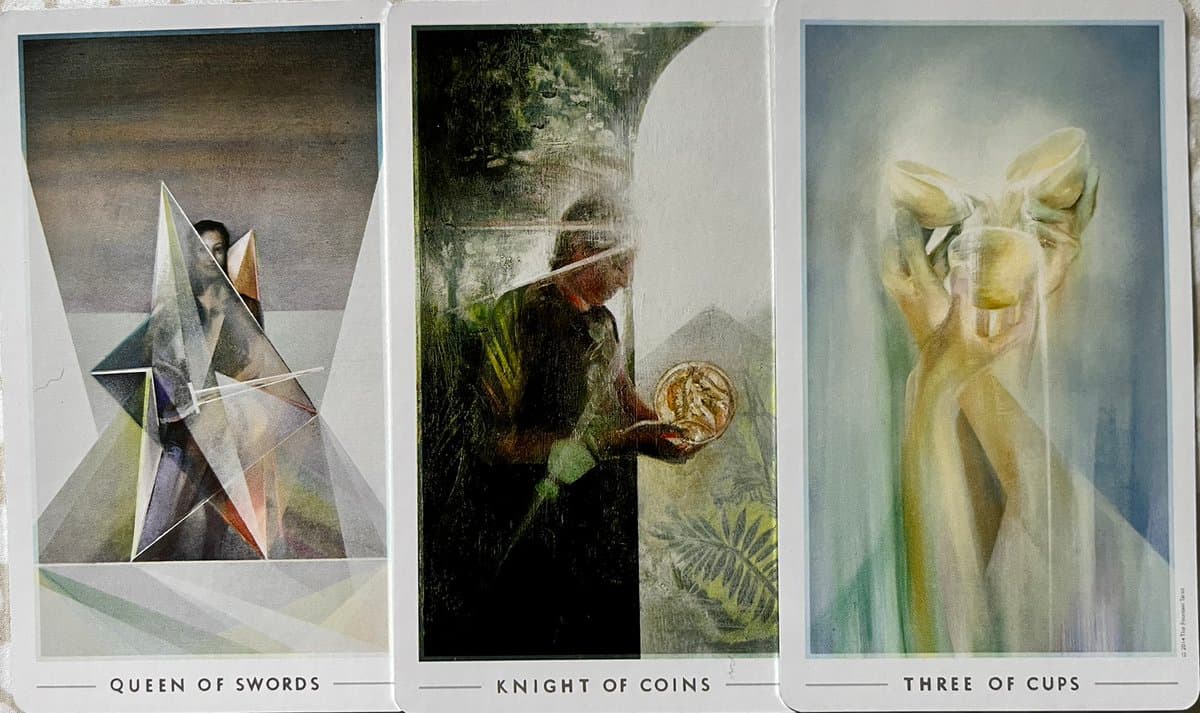 A photograph of three tarot cards laid side-by-side, featuring the Queen of Swords, Knight of Coins, and Three of Cups. The cards display artistic illustrations of figures and objects with their names printed at the bottom.