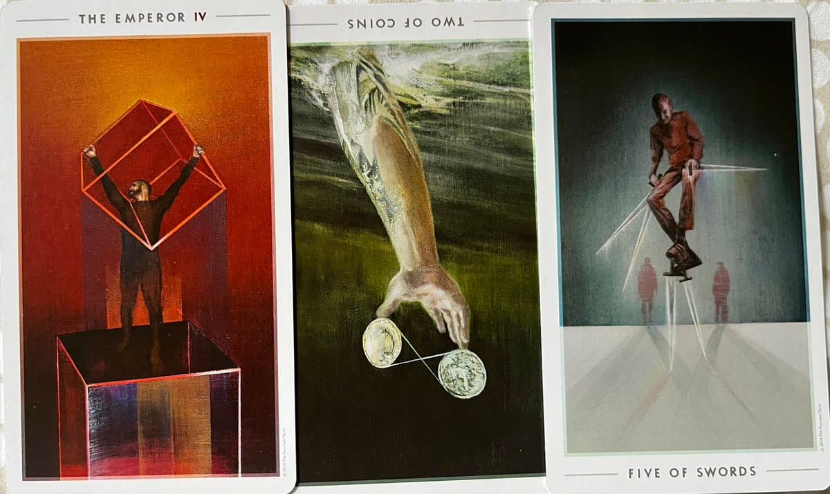 A photograph of three tarot cards laid side-by-side on a textured surface, featuring the Emperor, Two of Coins, and Five of Swords.