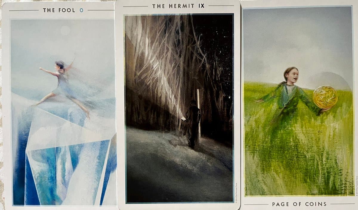A photograph showing three tarot cards laid side-by-side: The Fool, The Hermit, and Page of Coins. The cards feature watercolor-style illustrations of figures in symbolic settings.