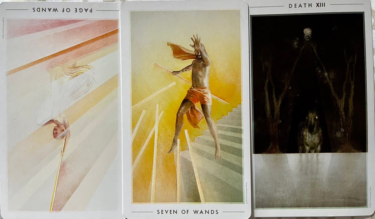 A photograph of three tarot cards arranged side-by-side, showing the Page of Wands (upside down), the Seven of Wands, and the Death card.