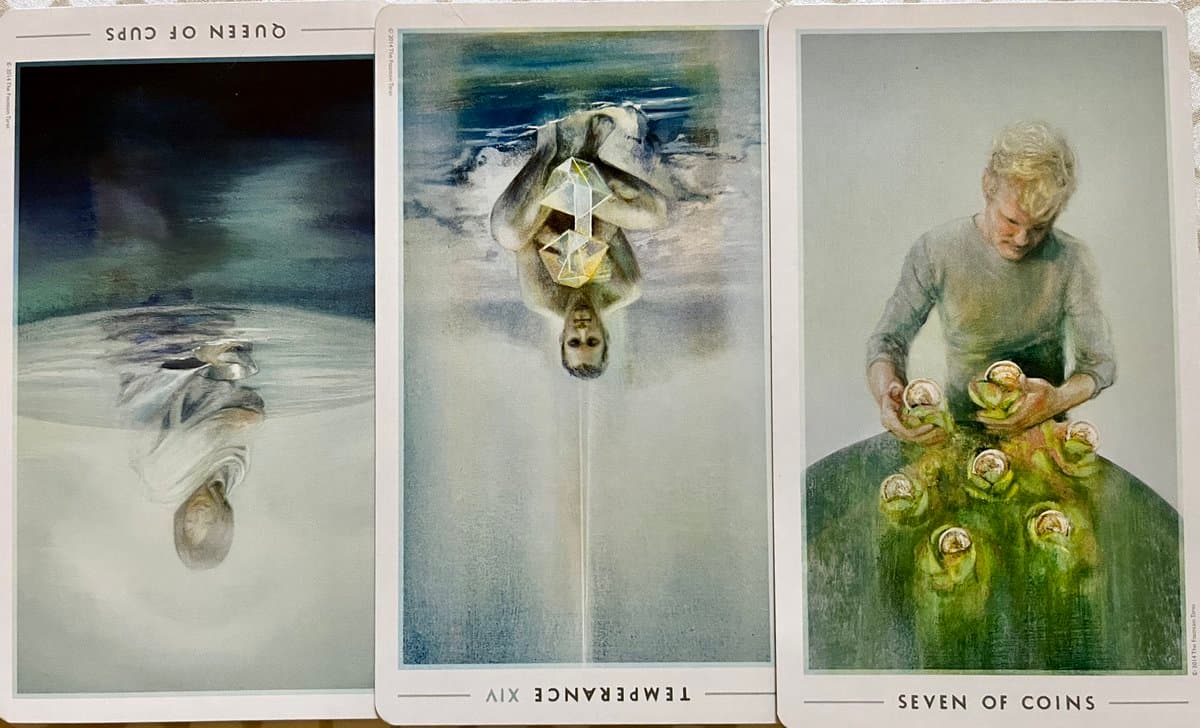 A photograph of three tarot cards laid side-by-side, showing the Queen of Cups, Temperance, and Seven of Coins. The first two cards are displayed upside down, while the third is right-side up.
