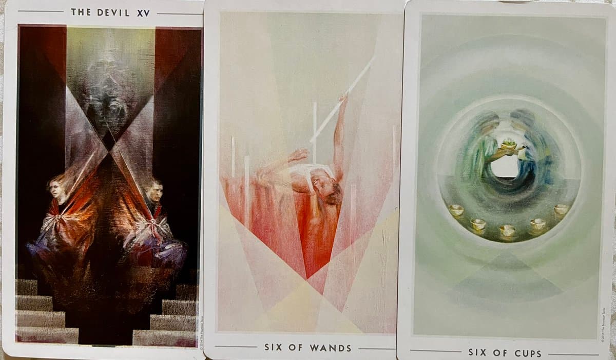 A photograph of three tarot cards laid side-by-side on a light surface. The cards feature artistic illustrations with a modern, geometric style.