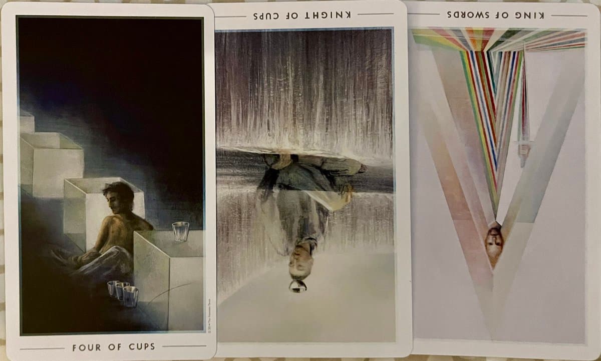 A photograph of three tarot cards laid out side-by-side, showing the Four of Cups, Knight of Cups, and King of Swords. The cards feature surreal illustrations of figures in abstract, geometric, or reflective settings.