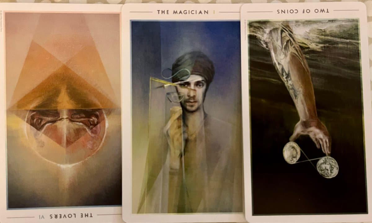 A photograph showing three tarot cards laid side-by-side on a patterned surface. The cards depict 'The Lovers' (upright), 'The Magician' (upright), and 'Two of Coins' (upside down).