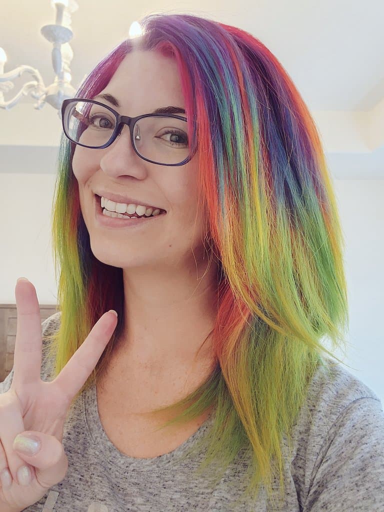 A portrait of a smiling woman with long, brightly dyed rainbow hair and glasses, making a peace sign with her hand.
