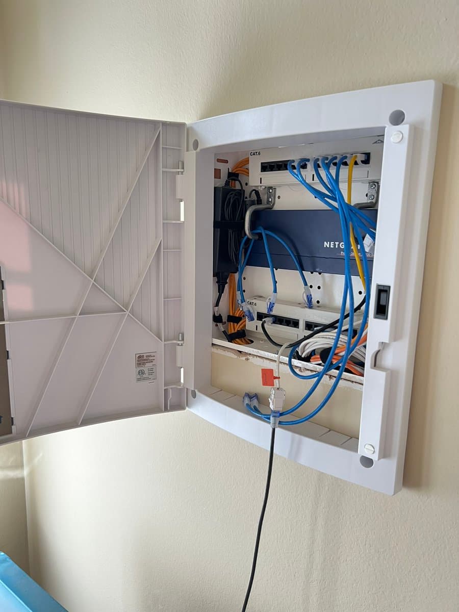 A wall-mounted white network patch panel enclosure is shown with its door open, revealing a dense and complex arrangement of blue and orange Ethernet cables connected to a Netgear switch and multiple wall jacks. The setup includes a power supply unit and various cable management ties, illustrating an extensive in-wall networking infrastructure.