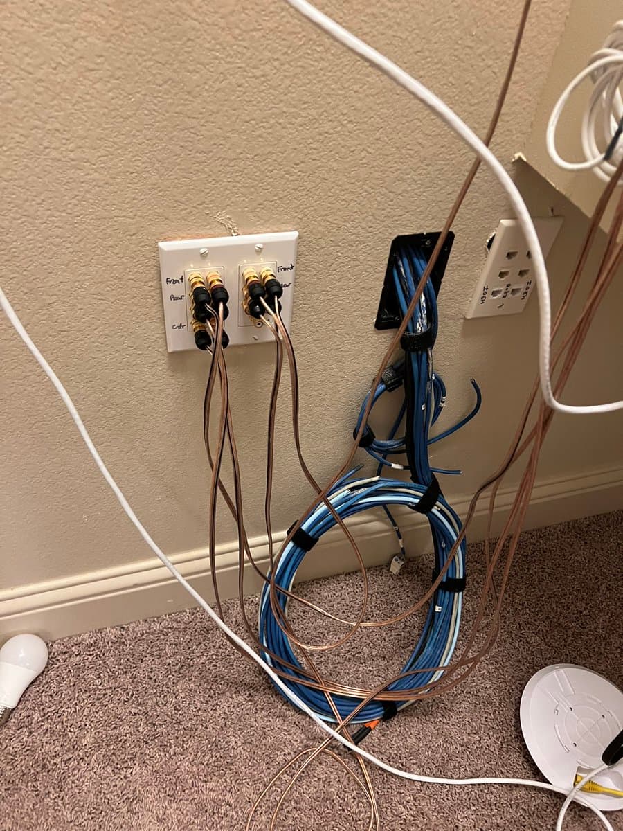 A photograph of a wall corner showing an extensive and cluttered network of cables, including coiled blue Ethernet cables and numerous coaxial cables plugged into wall jacks. The image highlights a dense setup of networking hardware, consistent with the context of a 'networking psycho'.