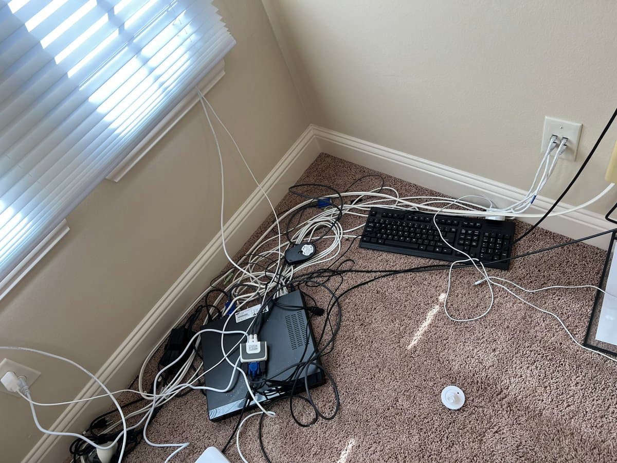 A photograph of a corner of a room with carpeted flooring, showing a computer tower and keyboard on the floor surrounded by a tangled mess of white and black cables. The wires run from the equipment to wall outlets and a window, illustrating a chaotic network setup.