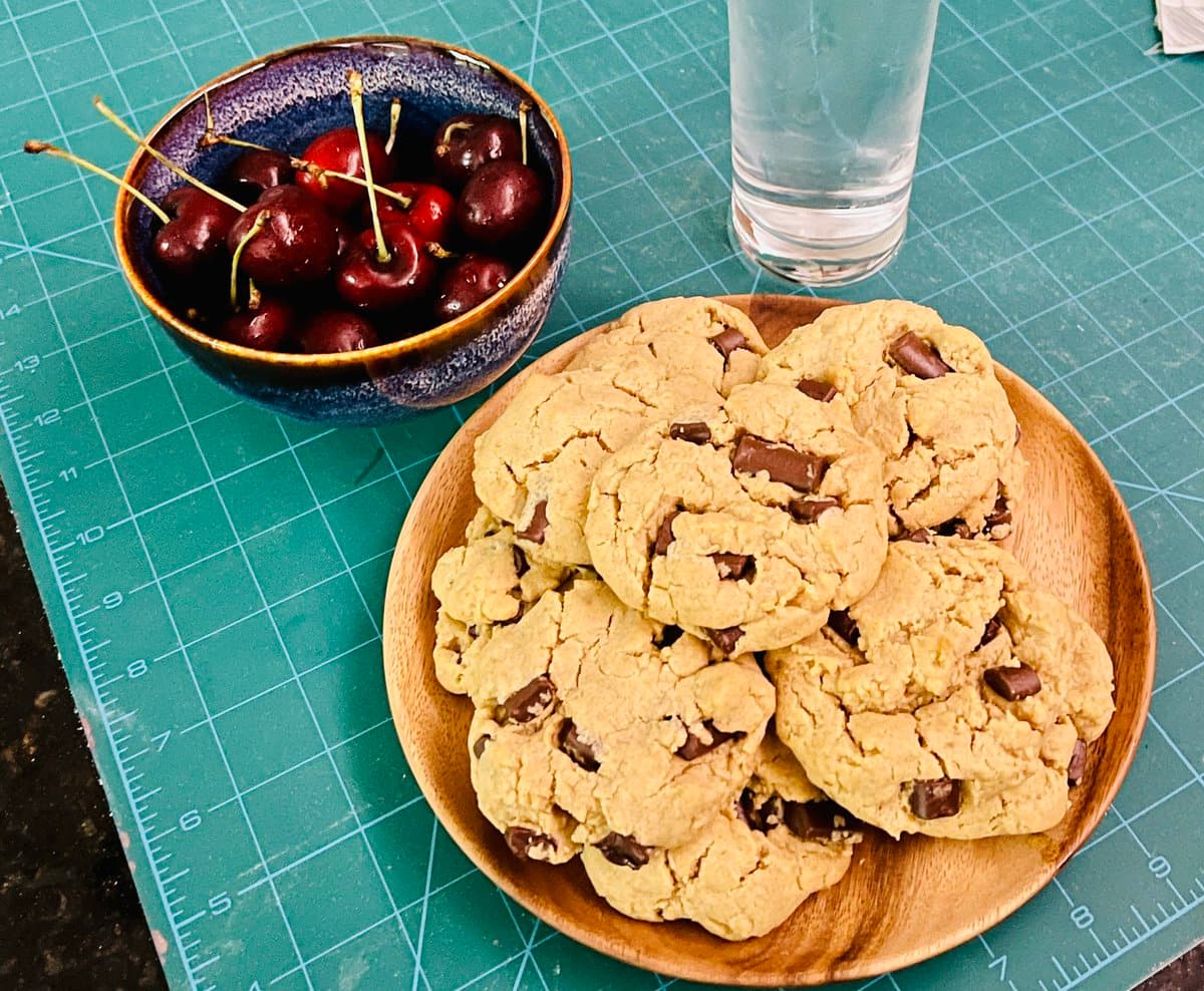 A top-down photograph of a plate of chocolate chip cookies and a bowl of fresh cherries sitting on a green cutting mat next to a glass of water.