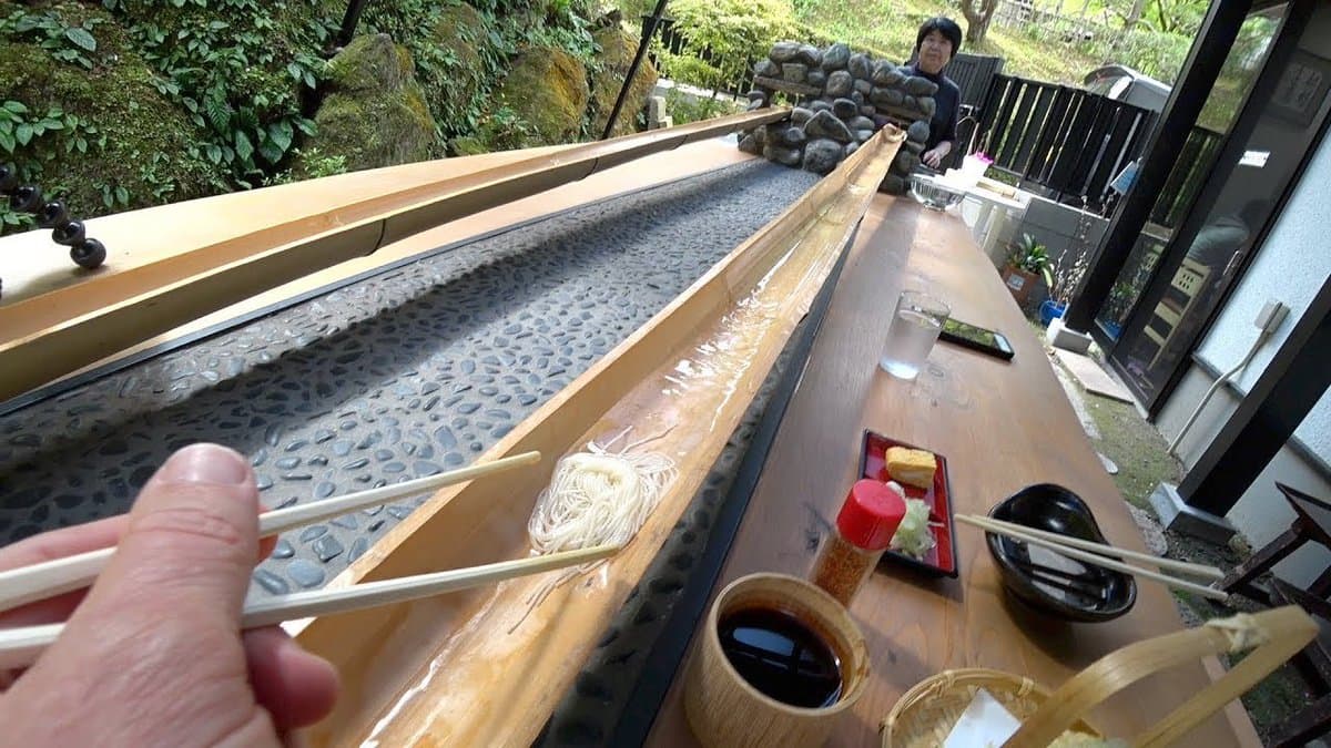 A first-person view of a person using chopsticks to pick up a bundle of noodles from a water-filled wooden trough at an outdoor noodle slide restaurant. The setting includes a long wooden counter with condiments and dishes, a stone-lined water channel, and a lush green garden in the background.