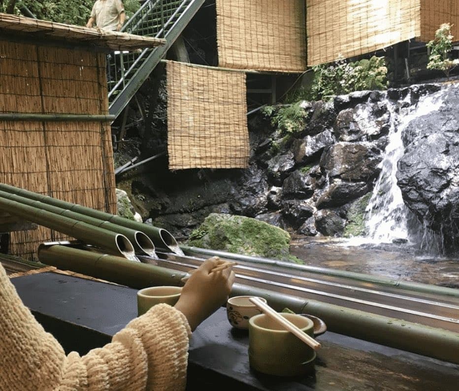 A person sits at a bamboo counter with bowls and chopsticks, watching water flow from bamboo pipes into a stream beside a small waterfall and bamboo-walled structures.