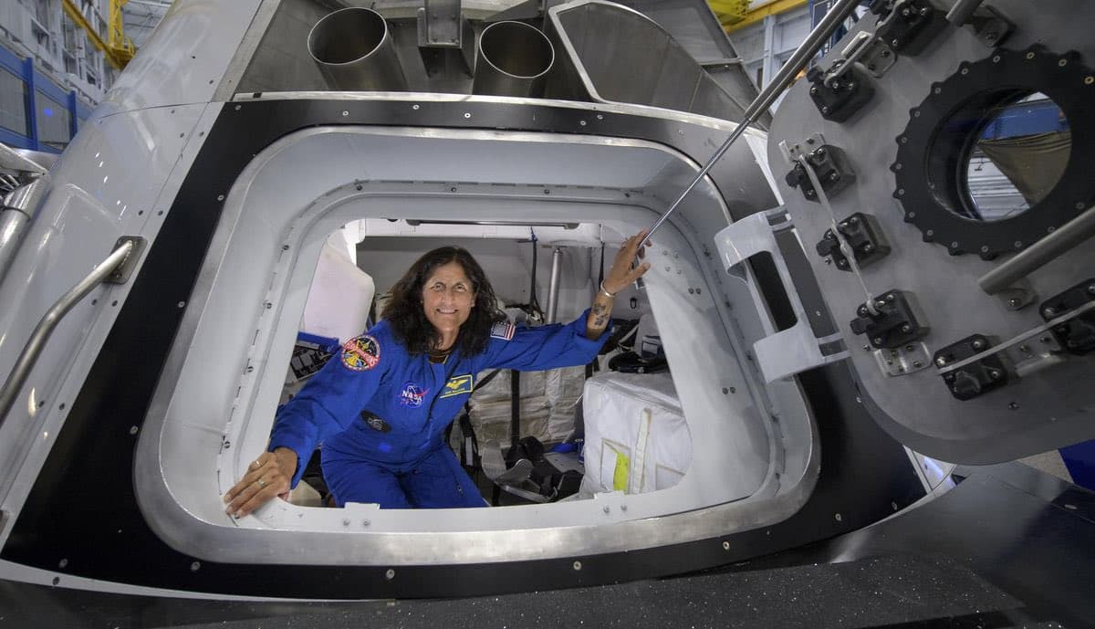 A woman in a blue NASA flight suit sits inside the open hatch of a spacecraft capsule, smiling and reaching toward the door mechanism. The image is a photograph taken inside a facility, likely a clean room or assembly area.