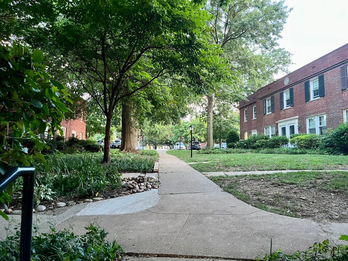 A photograph of a paved walking path winding through a landscaped area with trees and bushes, leading toward a large red brick apartment building with black shutters.